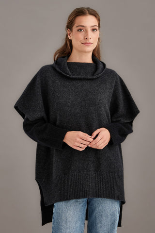 Woman wearing a dark gray poncho over a black sweater with blue jeans on a gray background