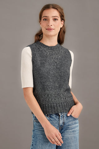 Woman wearing a gray knitted vest over a white shirt with blue jeans on a gray background