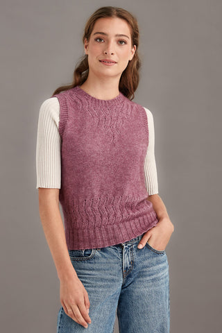 Woman wearing a heather pink knitted vest over a white shirt with blue jeans on a gray background