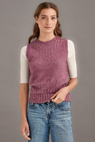 Woman wearing a heather pink knitted vest over a white shirt with blue jeans on a gray background