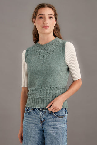 Woman wearing a mint green knitted vest over a white shirt with a gray background