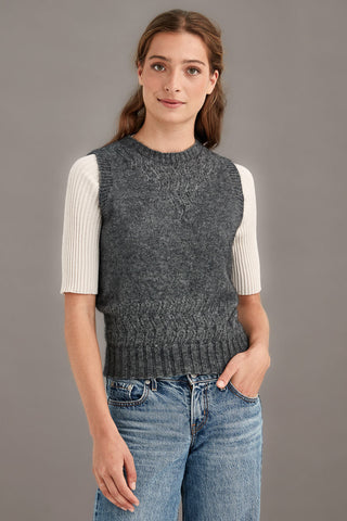 Woman wearing a gray knitted vest over a white shirt with blue jeans on a gray background