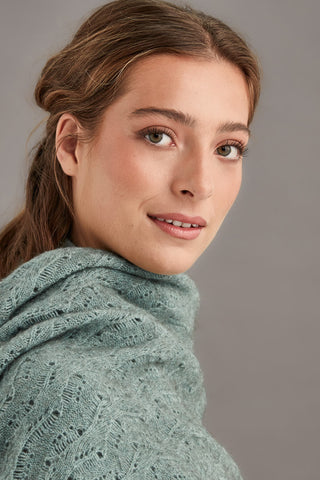 Woman wearing a mint green knitted shawl over a blue sweater against a gray background