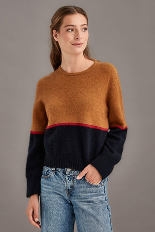 Woman wearing a colourblocked sweater with a brown top and black bottom with a red stripe paired with blue jeans against a gray background.
