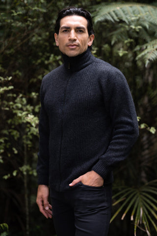 A man wearing a black rib front jacket with a turtleneck neckline, standing outdoors with foliage in the background.