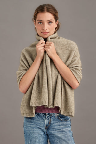 Woman wearing a beige travel throw over a maroon shirt with blue jeans against a gray background