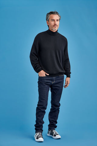 Man wearing a black turtleneck sweater, dark jeans, and black and white sneakers on a blue background