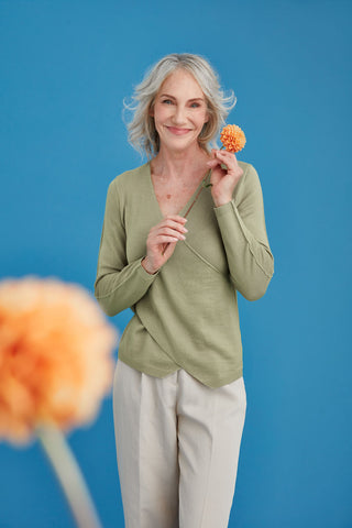 Woman wearing a citron merino top and holding an orange flower against a blue background